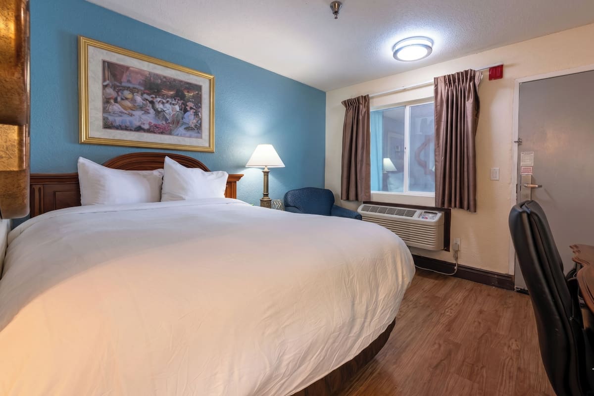 A hotel room with a large bed, a desk, a chair, a window with curtains, and a wall-mounted air conditioner.