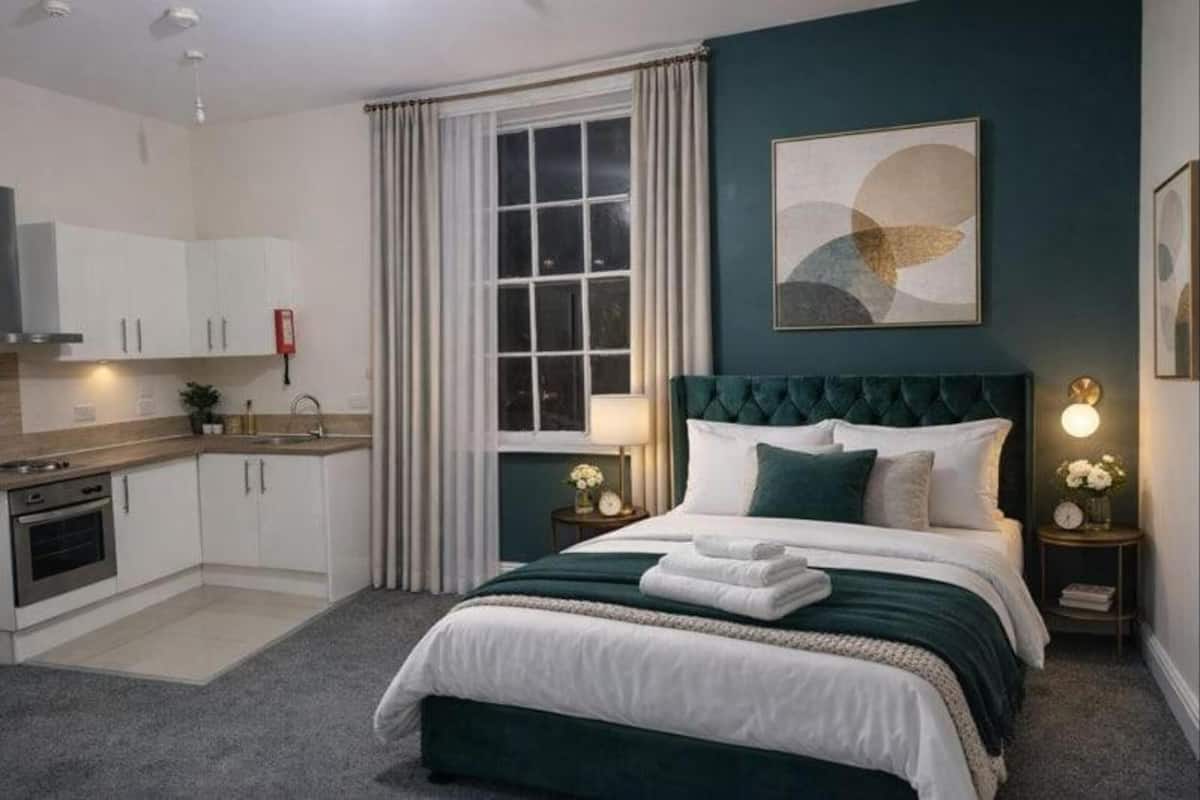 A bedroom with a bed, a nightstand, a lamp, a window, and a kitchen area.