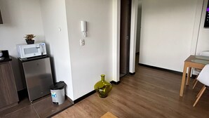 A modern, minimalistic living space with a wooden dining table, a small kitchen area with a microwave, a trash bin, and a potted plant.