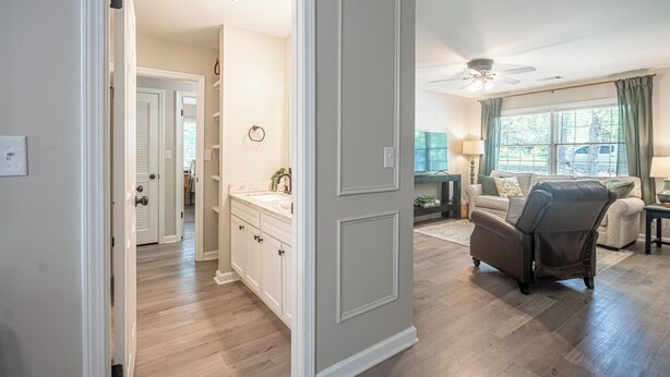 A well-lit hallway leading to a bathroom with a vanity, a ceiling fan, and a view of a living area with a sofa and a chair.