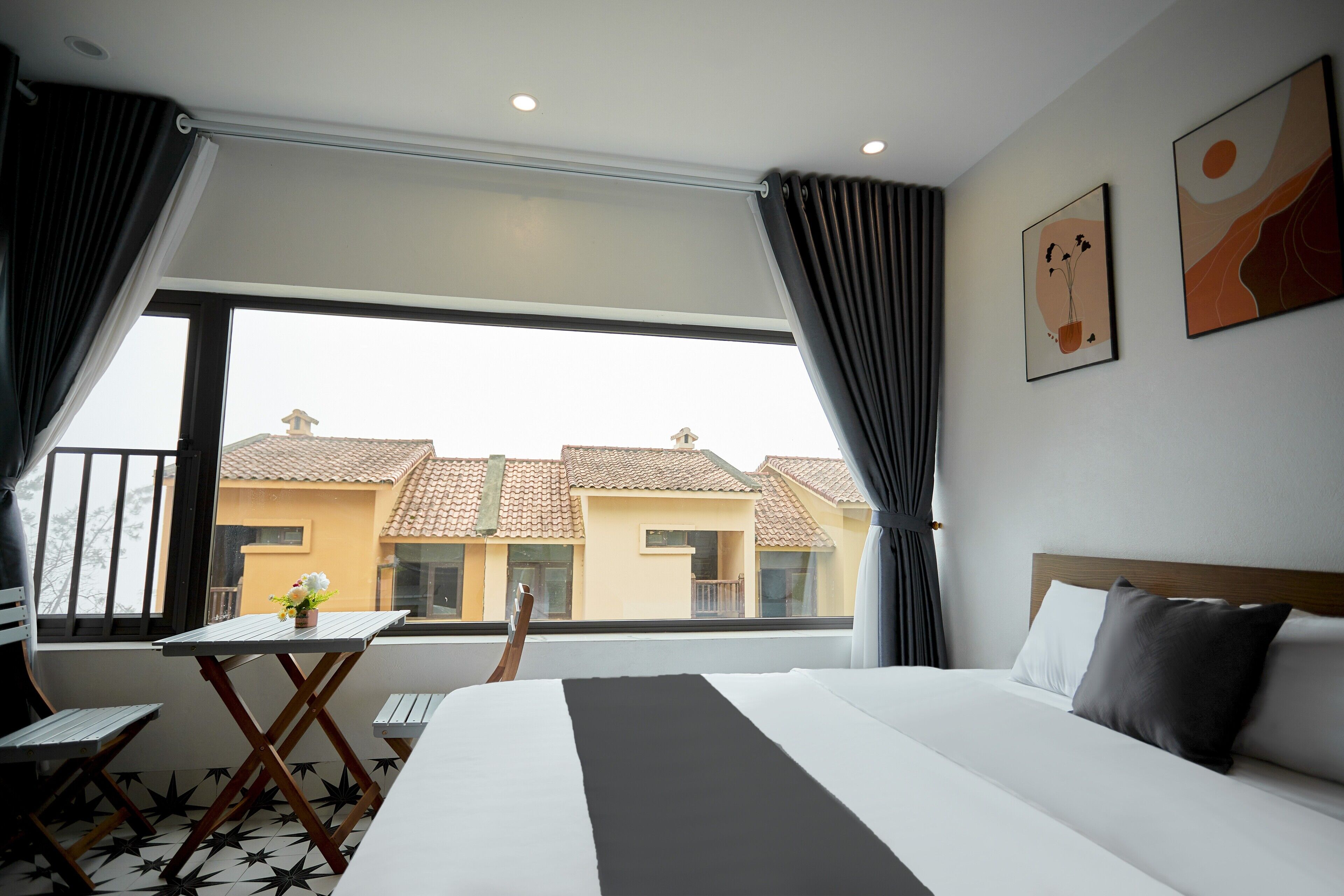 A hotel room with a bed, a small table with a flower, a chair, and a balcony with a view of buildings.