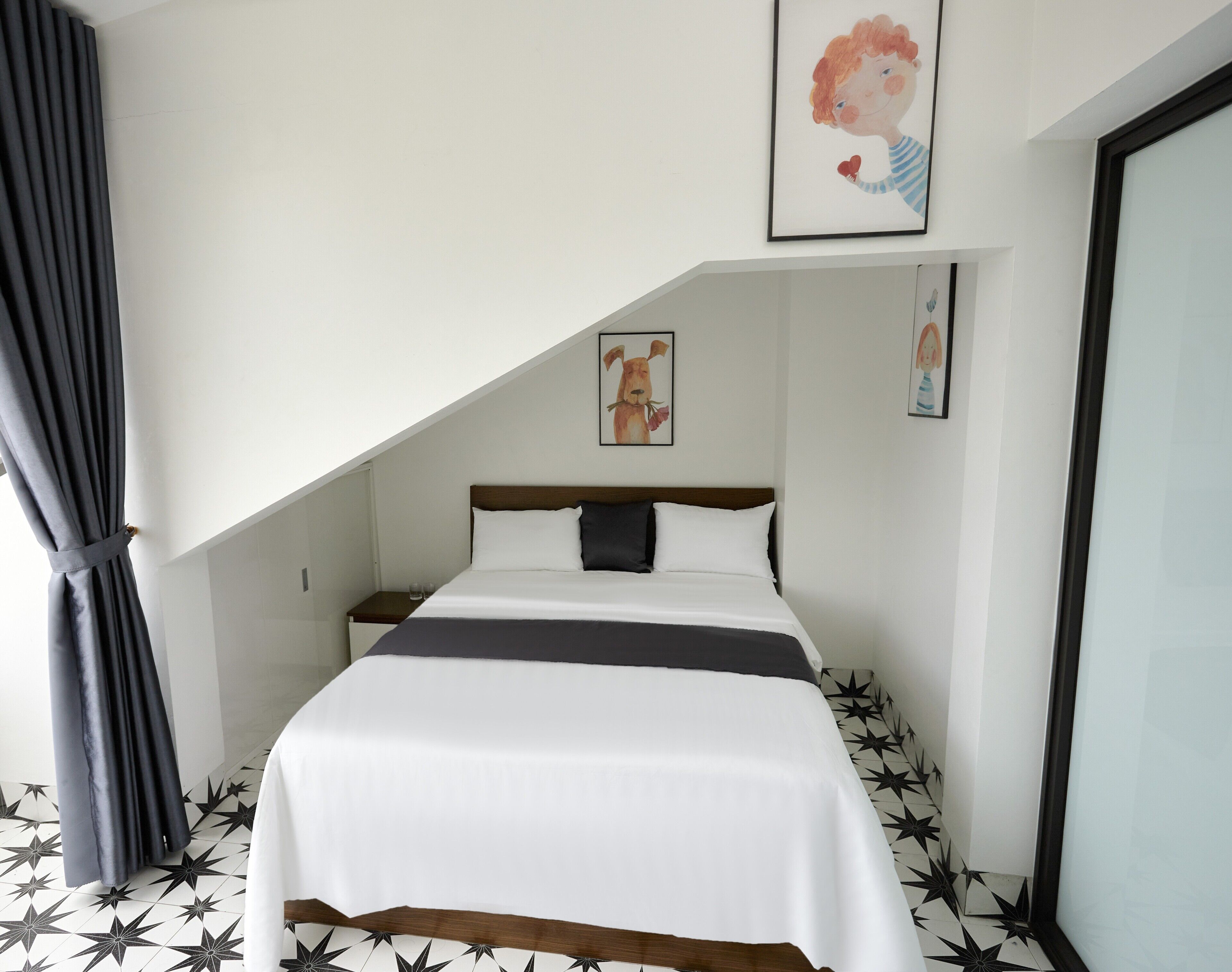 A bedroom with a bed, a nightstand, a window with curtains, and a wall with framed artwork.