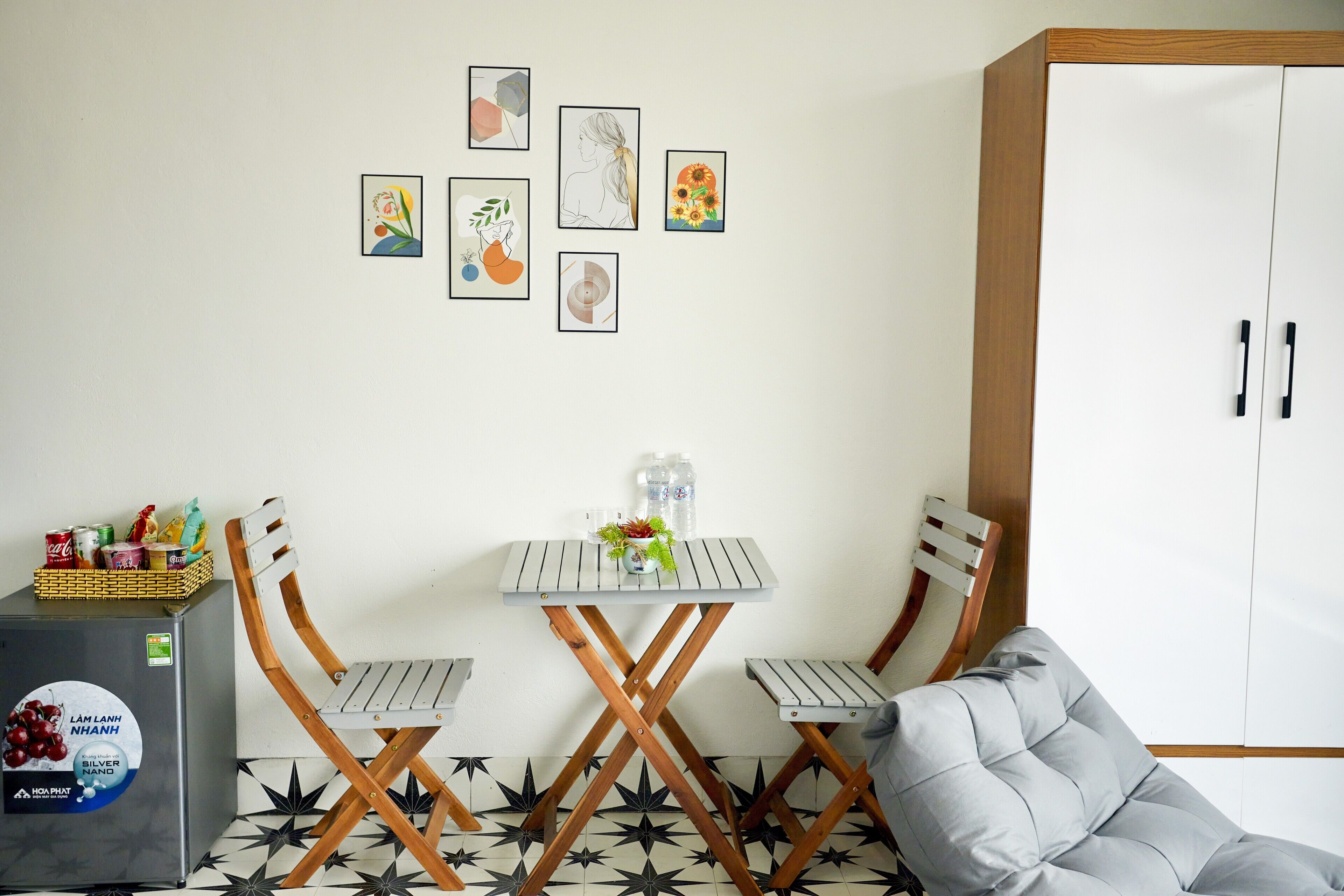 Habitación compacta con kitchenette, dos sillas plegables, una mesita pequeña y un sofá gris. Se observan obras de arte enmarcadas en la pared y un armario con pomos negros.