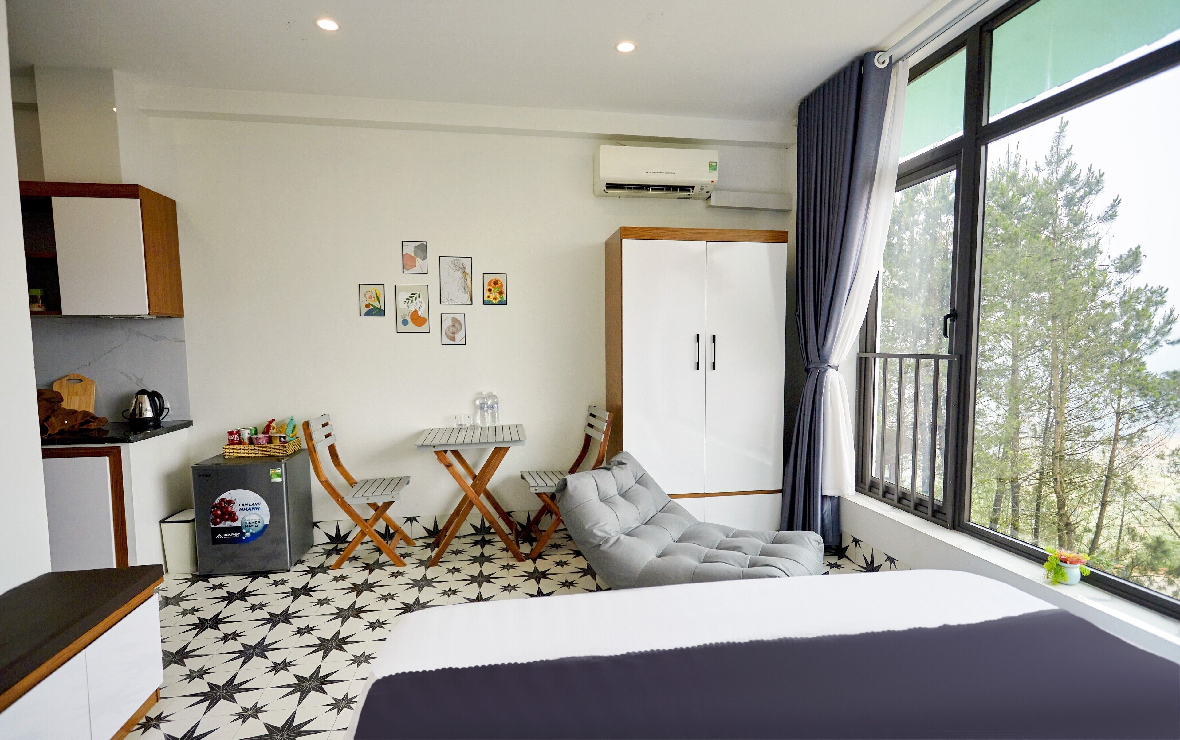Ein Hotelzimmer mit Bett, Schreibtisch mit Stühlen, Kleiderschrank und großem Fenster mit Blick auf Bäume.