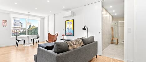 A modern living room with a grey sofa, a dining table with chairs, a painting on the wall, and a view of the city through large windows.