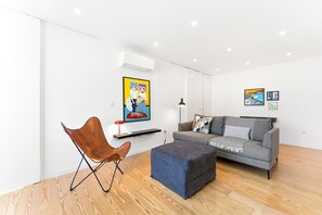 Ein modernes Wohnzimmer mit einem grauen Sofa, einem braunen Ledersessel und einem blauen Hocker. Ein Flachbildfernseher ist an der Wand montiert, und eine Stehlampe ist sichtbar.