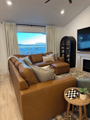 A modern living room with a large sectional sofa, a flat-screen TV, a fireplace, and a view of a distant landscape.
