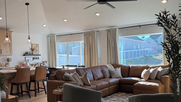 A spacious living room with a large brown leather sofa, a ceiling fan, and a dining area with wooden furniture. - Cozy Winter Escape, New Build with 3 Car Garage (Tooele)