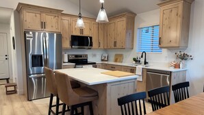 A modern kitchen with wooden cabinets, stainless steel appliances, a central island with stools, and pendant lighting. - Cozy Winter Escape, New Build with 3 Car Garage (Tooele)