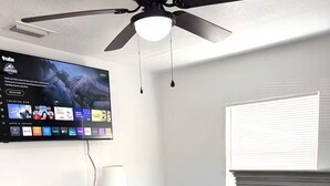 Ein Schlafzimmer mit an der Wand montiertem Fernseher, einem Schminktisch und einem Bett mit grauer Bettwäsche sowie einem Deckenventilator.