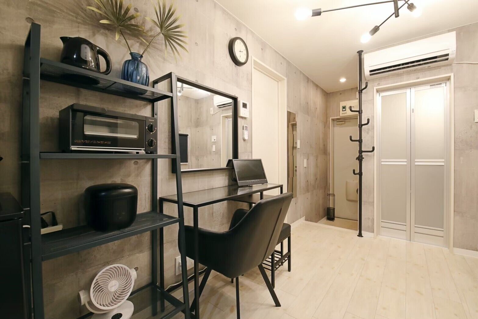 A modern kitchen with a microwave, kettle, and fan. There is a laptop on a desk and a chair. The room has wooden flooring and a clock on the wall.