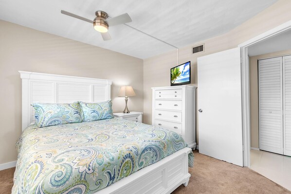 A bedroom with a bed, a nightstand, a dresser, a ceiling fan, a TV, and a door. - Bayshore Yacht & Tennis 211 - Premier (Indian Shores)