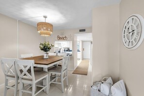 A dining area with a wooden table and white chairs, a hanging wicker light fixture, and a wall clock. - Bayshore Yacht & Tennis 211 - Premier (Indian Shores)
