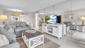 Una sala de estar moderna con televisor, muebles blancos y un espejo grande que refleja la habitación.