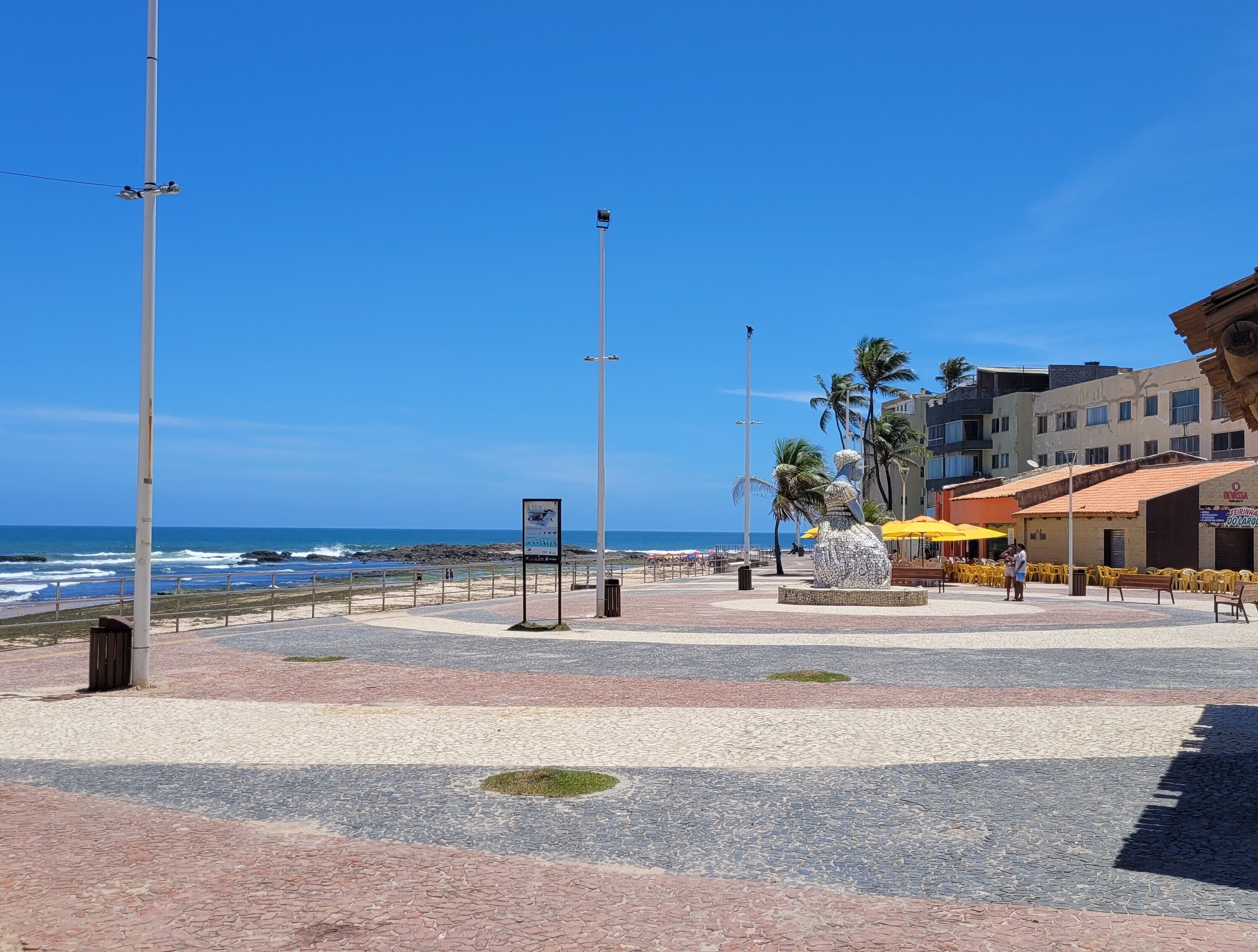 A coastal promenade with a paved walkway, palm trees, and a view of the ocean.