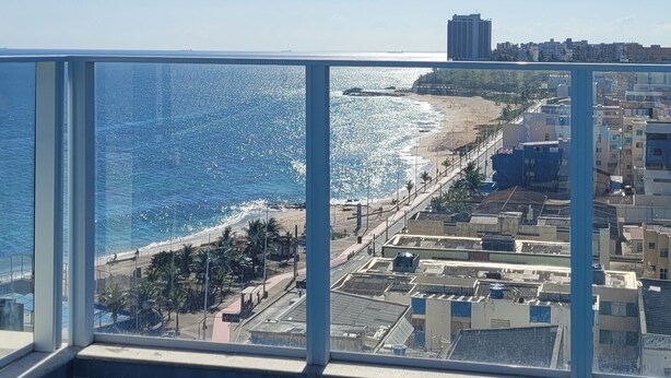 Uitzicht vanaf een balkon op een strand, met heldere lucht, een zwembad en gebouwen op de achtergrond.