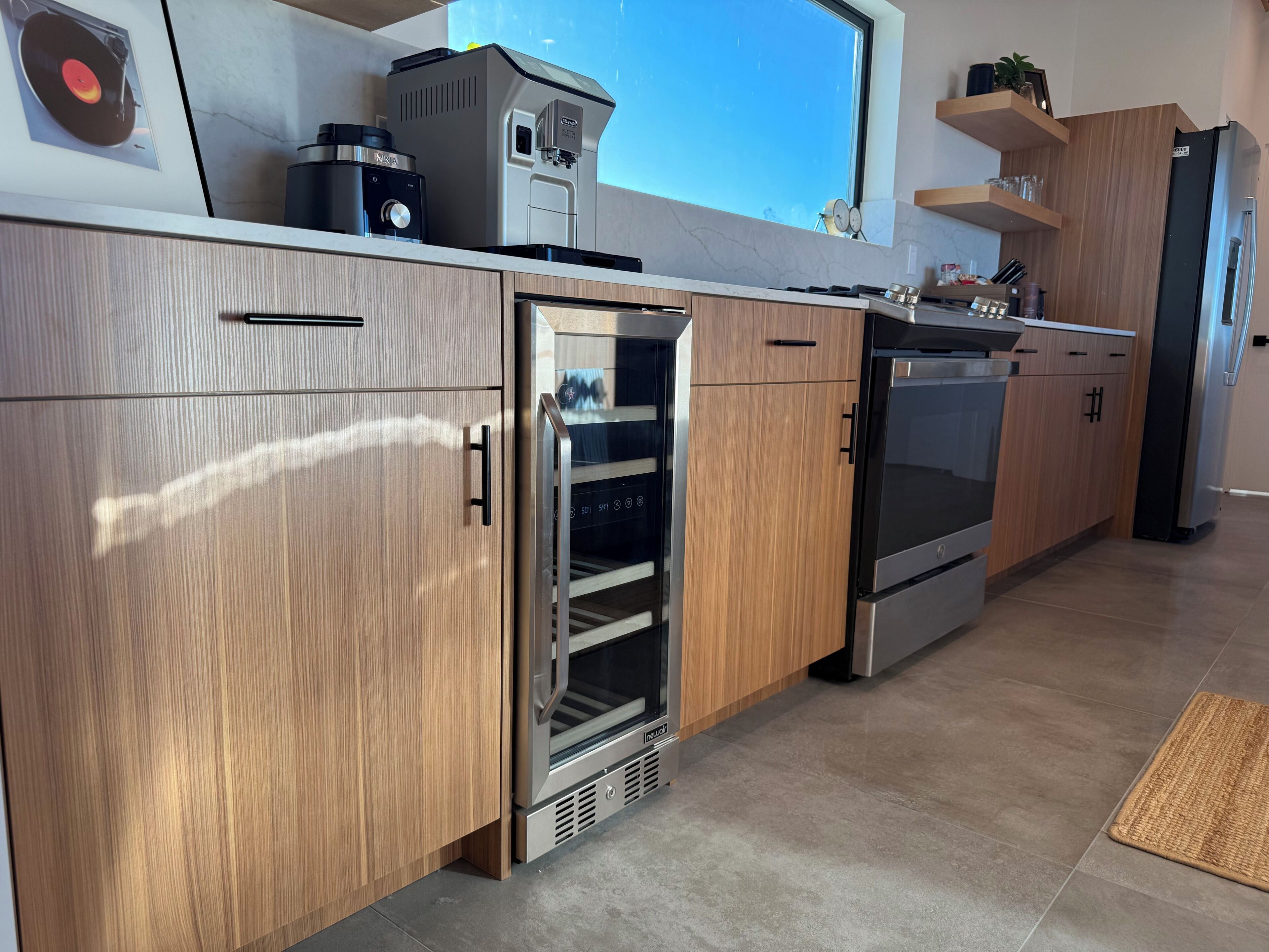 A modern kitchen with wooden cabinets, a built-in oven, a coffee machine, and a window with a view.