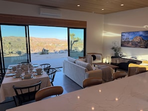 Ein Essbereich mit Tisch für vier Personen, einer Couch und einem Sessel, mit Blick auf eine Wüstenlandschaft durch große Fenster.