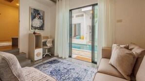 Ett modernt vardagsrum med en beige soffa, en mönstrad matta, en tavla på väggen och en glasdörr som leder ut till ett poolområde.