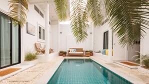 En swimmingpool med ett kaklat botten, omgiven av en vit vägg, palmer och en soffa med kuddar.