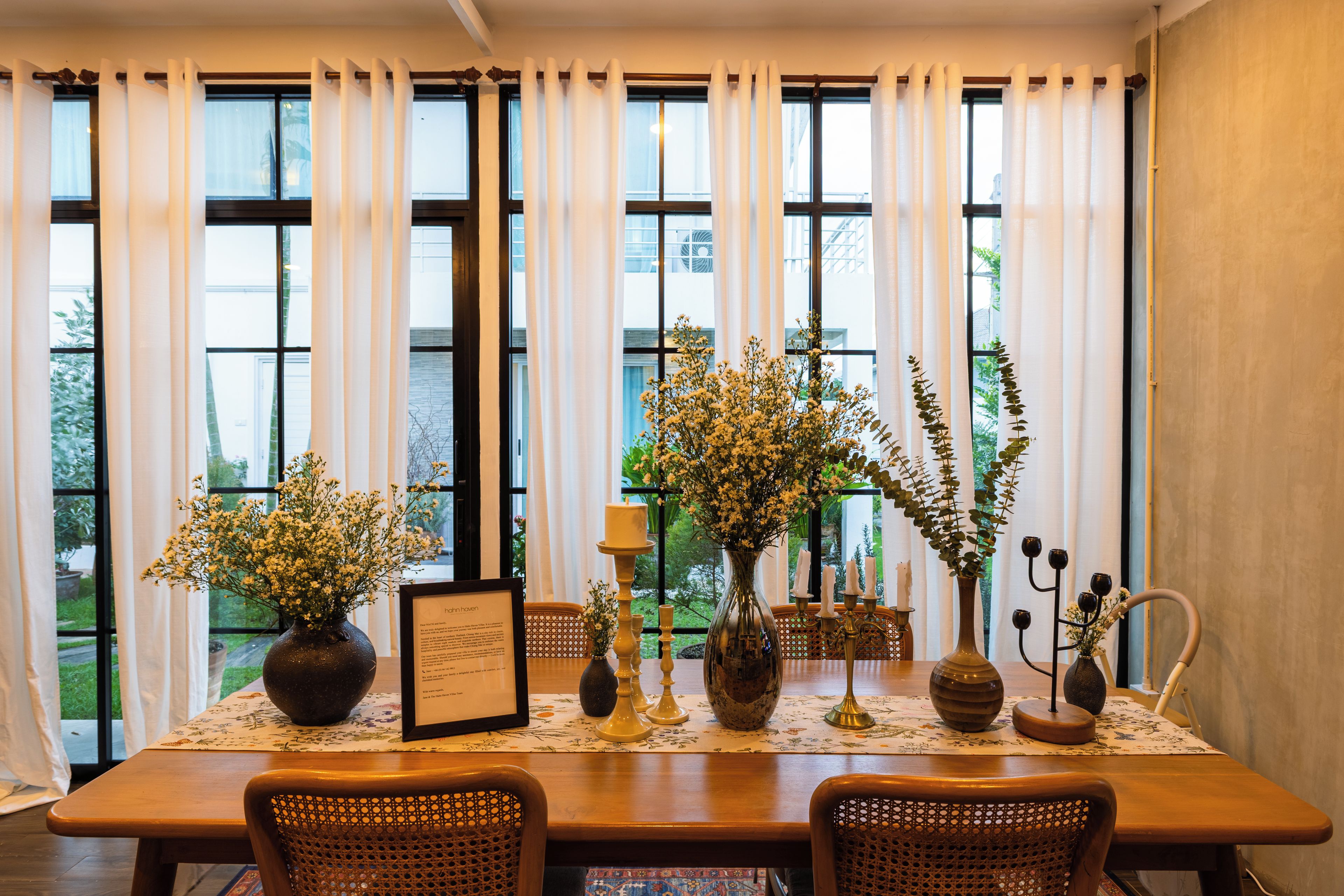 Un coin repas avec une table dressée pour deux personnes, entouré de grandes fenêtres ornées de rideaux blancs, et agrémenté de divers objets décoratifs, notamment des vases et des bougies.
