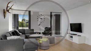 A modern living room with a grey sofa, ottomans, a round coffee table, a black armchair, a wall clock, and a flat-screen TV. - Stunning home in Hemmet with sauna (Hemmet)