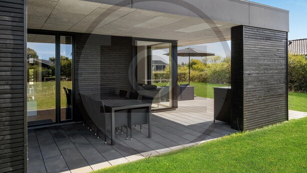 A modern outdoor patio with a dining table and chairs, surrounded by a wooden deck and glass doors leading to a garden. - Stunning home in Hemmet with sauna (Hemmet)