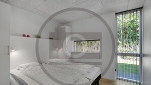 A modern bedroom with a large bed, a window with a view of greenery, and a small shelf with decorative items. - Stunning home in Hemmet with sauna (Hemmet)
