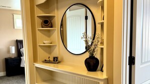 A hallway with a white console table, a round mirror, and a vase with flowers. Shelves are present on the console table. - Baldwin Retreat | Spacious Stay w/ 3 King Beds (Meridian)
