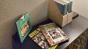 Eine gerahmte Karte mit Koordinaten, ein Schreibtisch mit Lampe, ein Stapel Spiralnotizbücher und zwei Bücher mit den Titeln 'Civil War' und 'Year of the Duck'.