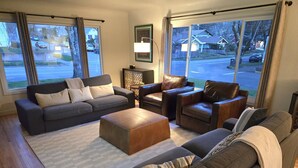 Ein modernes Wohnzimmer mit einem grauen Sofa, einem braunen Ledersessel und einem Holzfußhocker. An der Wand hängt ein gerahmtes Bild und auf einem Beistelltisch steht eine Lampe.