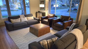 A modern living room with a grey sofa, a brown leather armchair, and a wooden ottoman. There is a framed picture on the wall and a lamp on a side table.