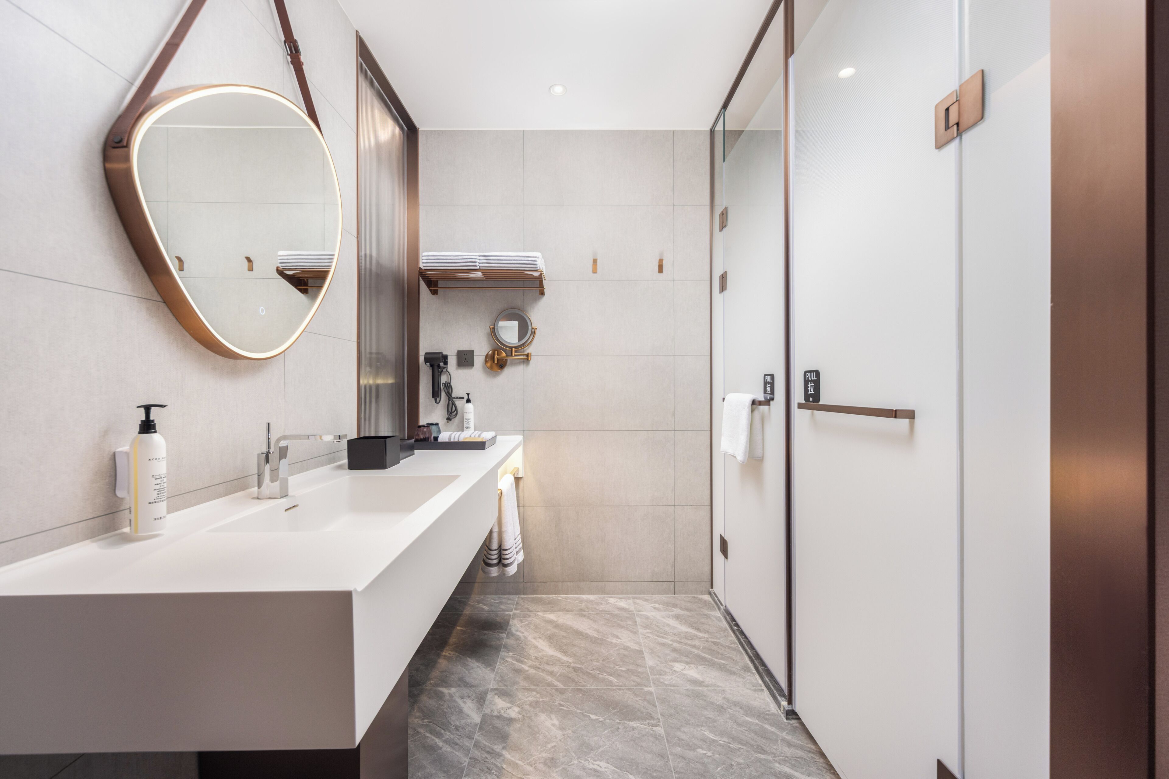 A modern bathroom with a white vanity, a round mirror, a towel rack, and a shower area.