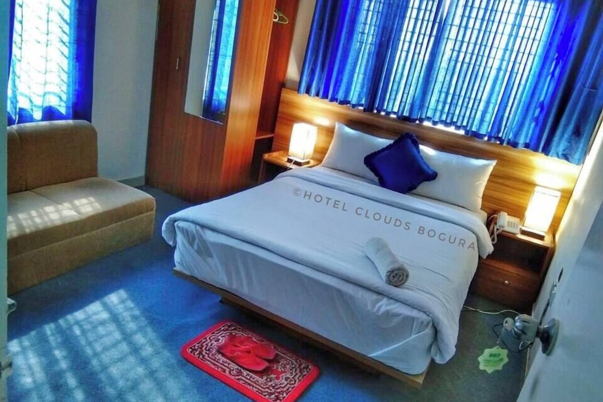 Een hotelkamer met een bed, een stoel, een nachtkastje met een lamp en een raam met blauwe gordijnen.