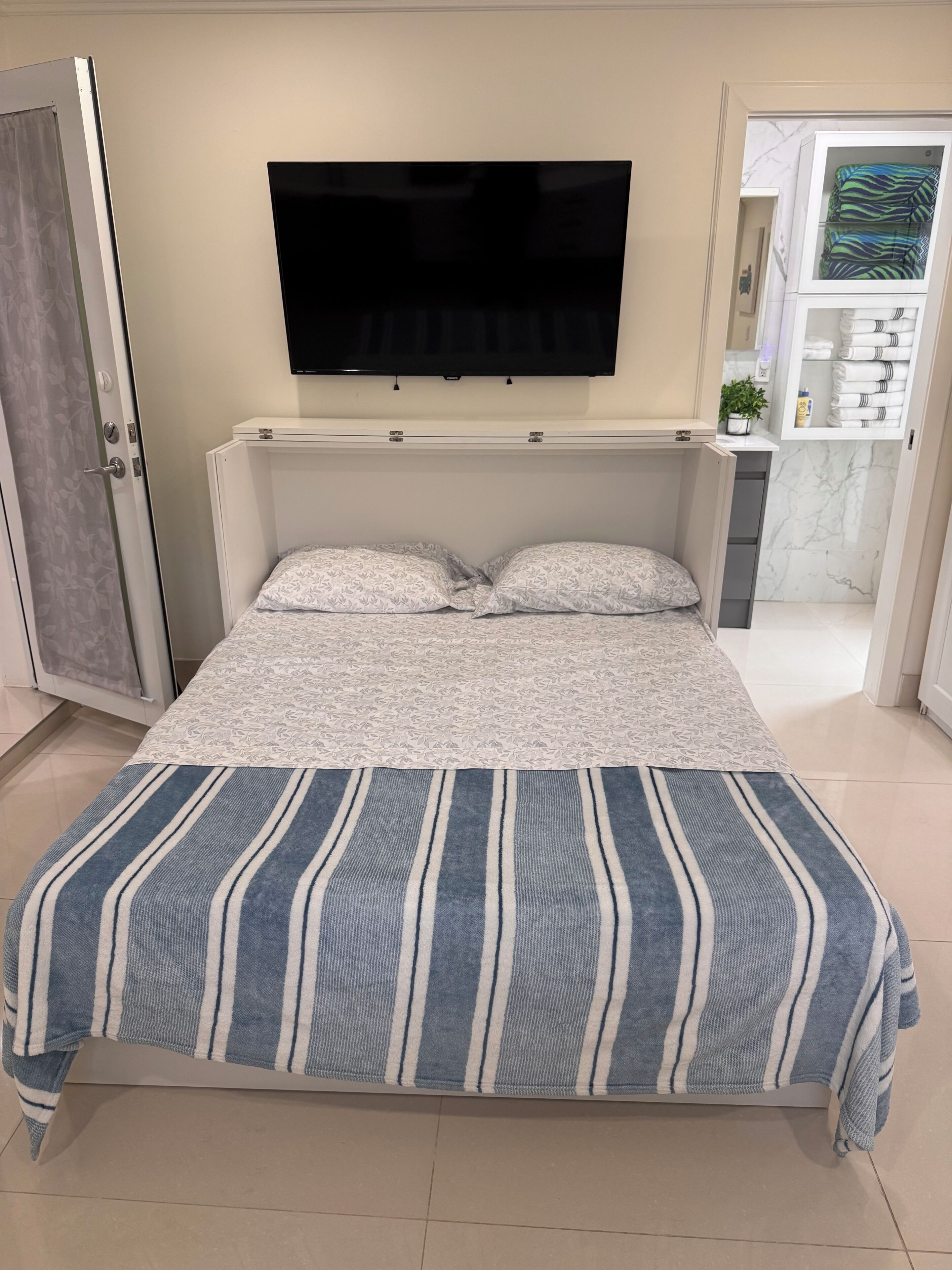 Une chambre à coucher avec un lit, un téléviseur fixé au mur et une salle de bain visible à travers une porte ouverte.
