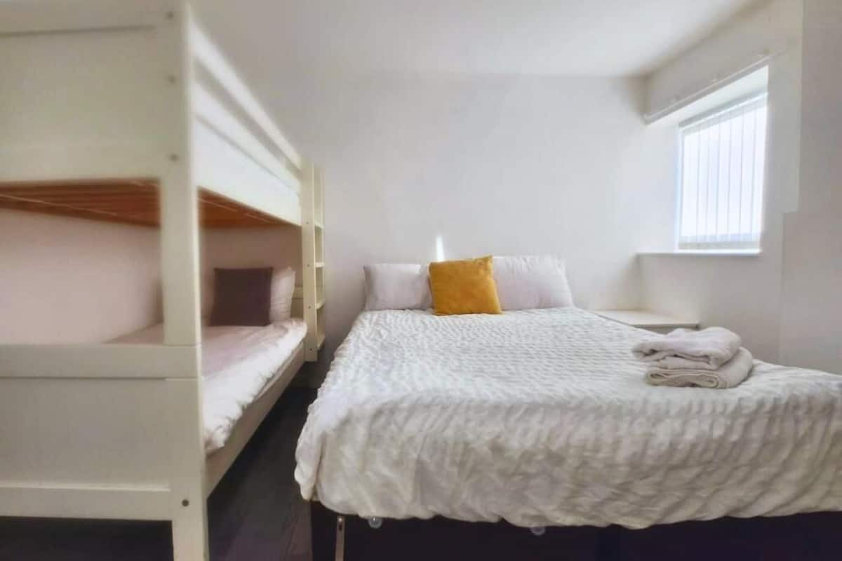 Un dormitorio con literas, una cama individual con colcha blanca, un almohadón amarillo y una ventana con persianas.