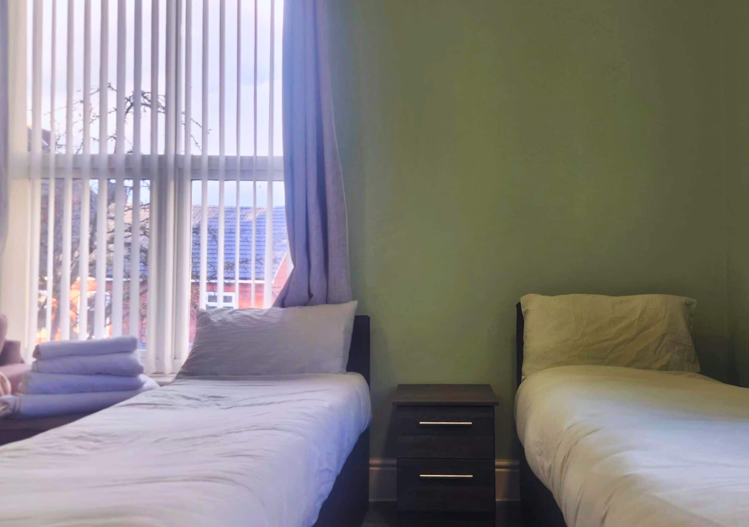 A hotel room with two beds, a window with blinds, and a view of buildings outside.