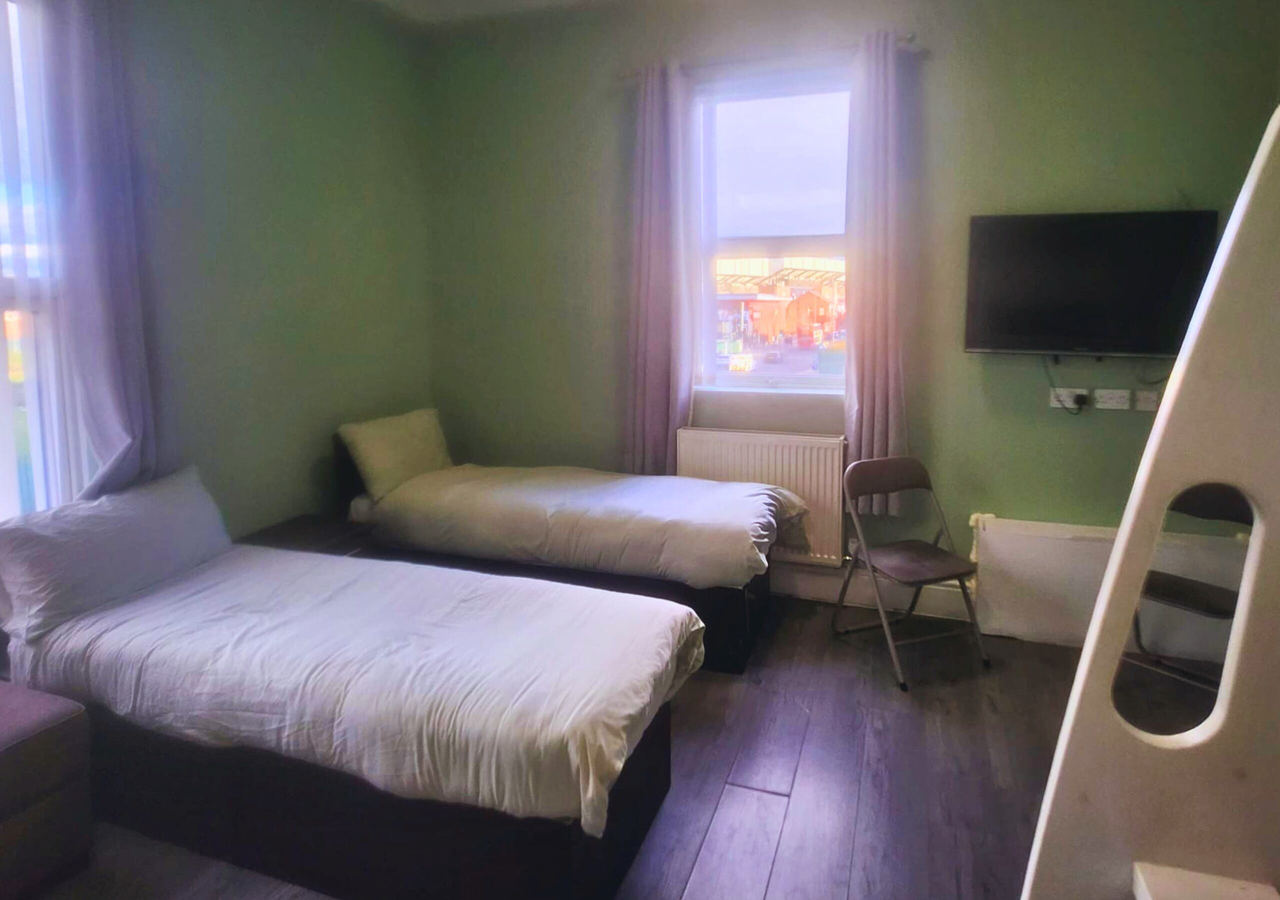 Una habitación con dos camas, una silla, un radiador, una ventana con cortinas y un televisor en la pared.