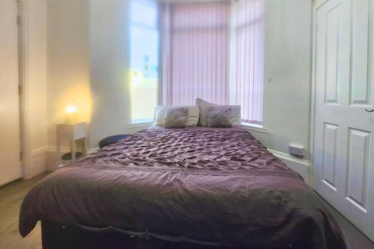 Un dormitorio con cama, almohadas, una mesita de noche y una ventana con persianas.