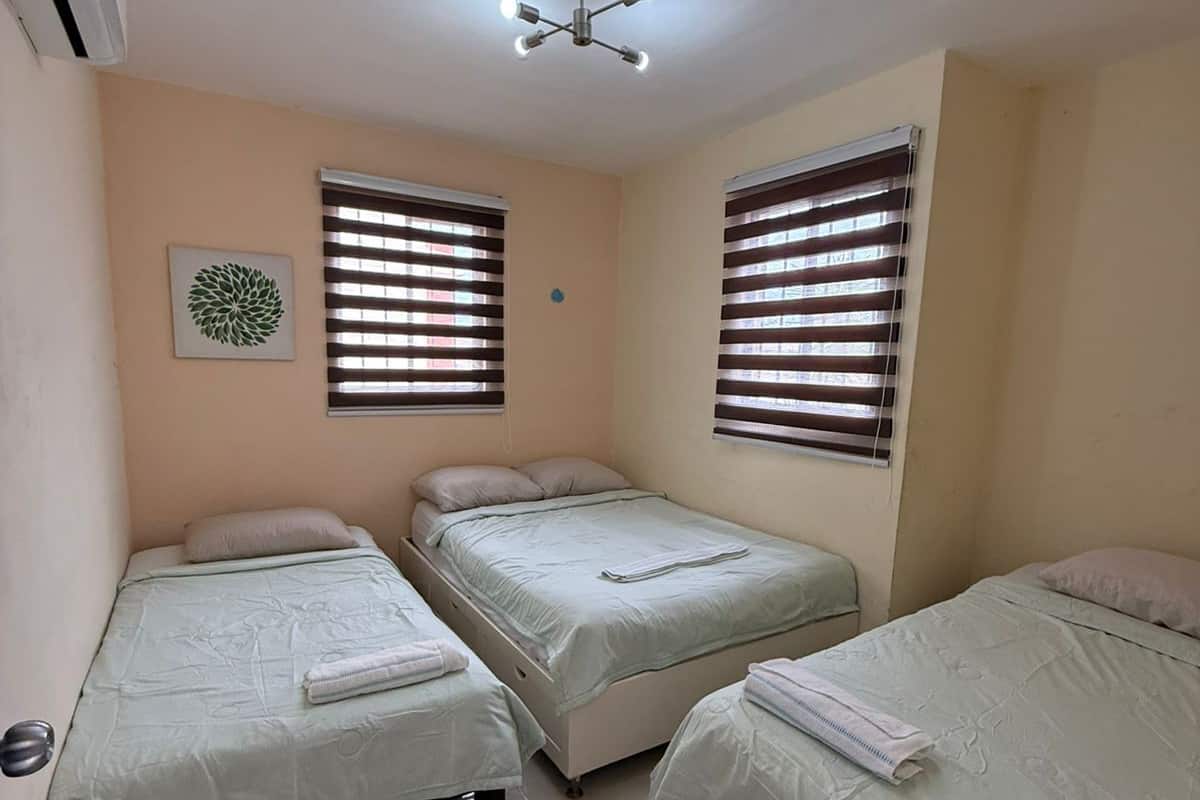 A room with three single beds, a ceiling fan, and two windows with blinds.