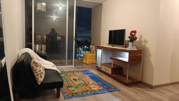 Een woonkamer met een zwarte bank, een kleurrijk vloerkleed, een televisie op een houten dressoir en een uitzicht op de stad 's nachts door een glazen deur.