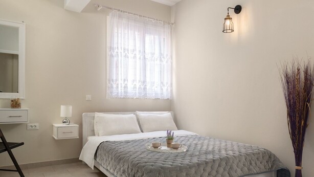 Une chambre à coucher comprenant un lit, une table de chevet, une lampe, une fenêtre avec des rideaux légers et un luminaire fixé au mur.