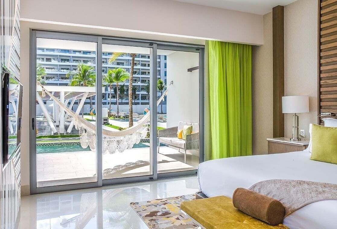 Una habitación de hotel con cama, un ventanal con vistas a una piscina y palmeras, una cortina verde y una lámpara en una mesita de noche.