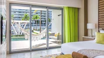 Una habitación de hotel con cama, un ventanal con vistas a una piscina y palmeras, una cortina verde y una lámpara en una mesita de noche.