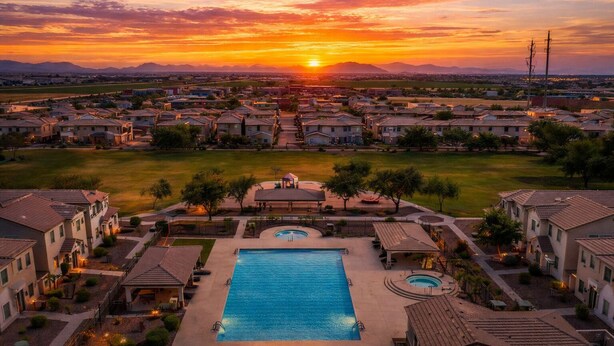 A residential area with a large swimming pool and a gazebo, surrounded by houses and a sunset view.