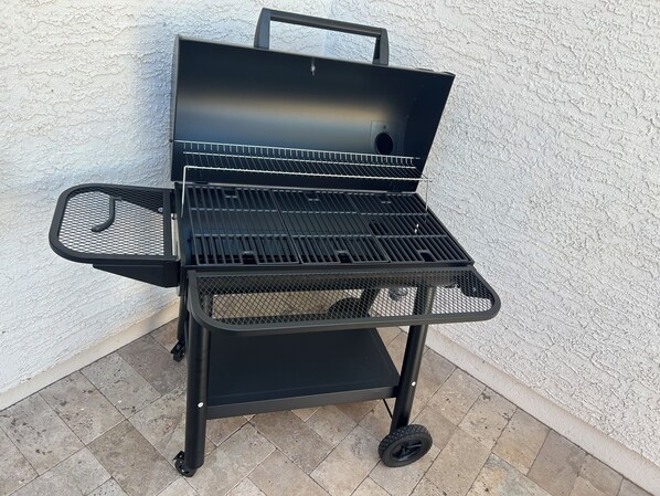 A black outdoor grill with a mesh lid, a handle, and a side shelf, set against a textured wall on a tiled patio.