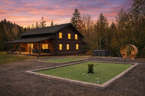 A log cabin with a porch and outdoor seating, surrounded by a manicured lawn and a putting green.