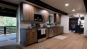A modern kitchen with wooden cabinets, stainless steel appliances, and a dining area with blue chairs. - Rainier Timber Retreat • Hot Tub & Sauna • Forest Deck on 5 Acres (Wilkeson)