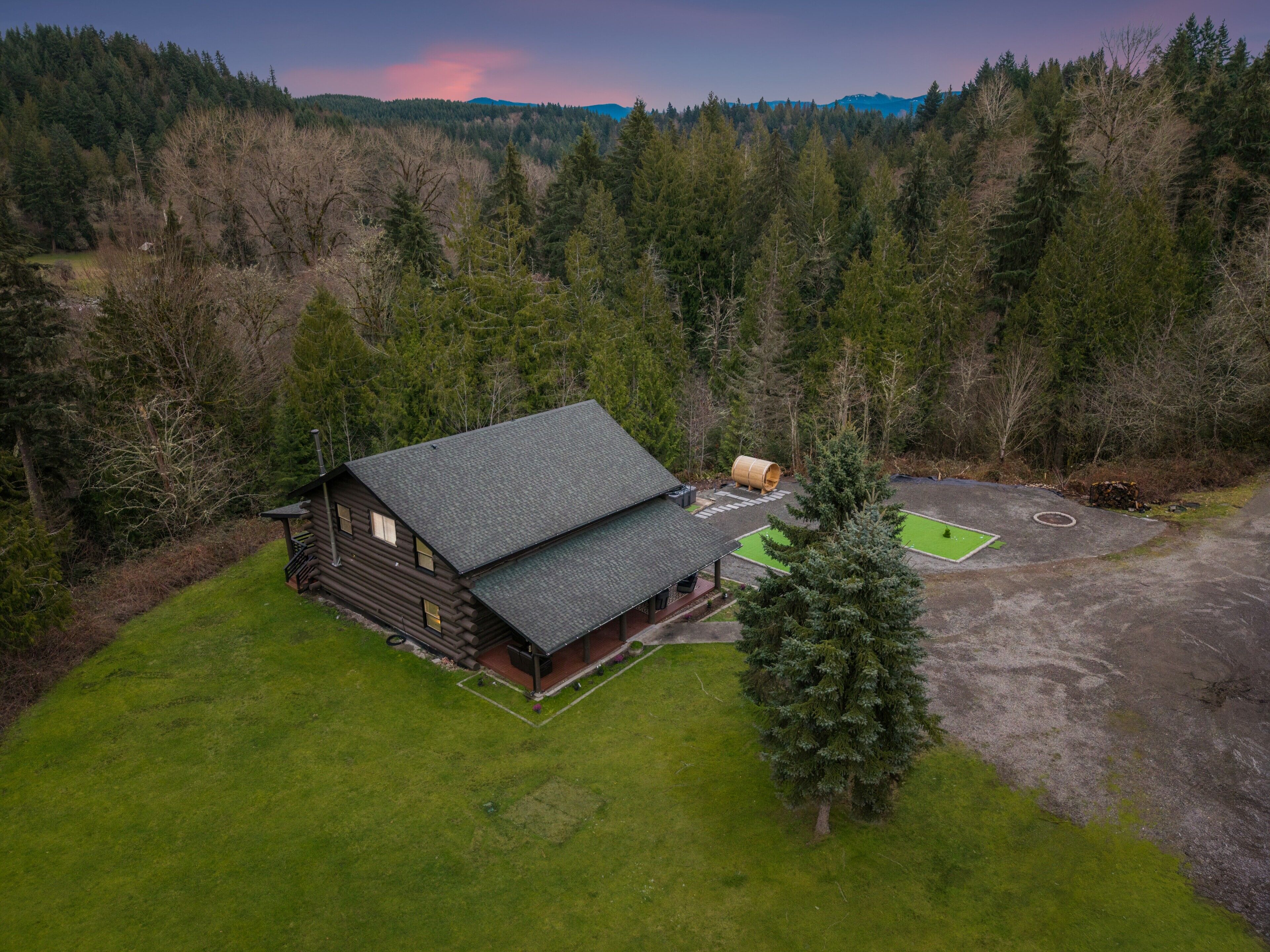 A log cabin with a deck and a green area, surrounded by forest.