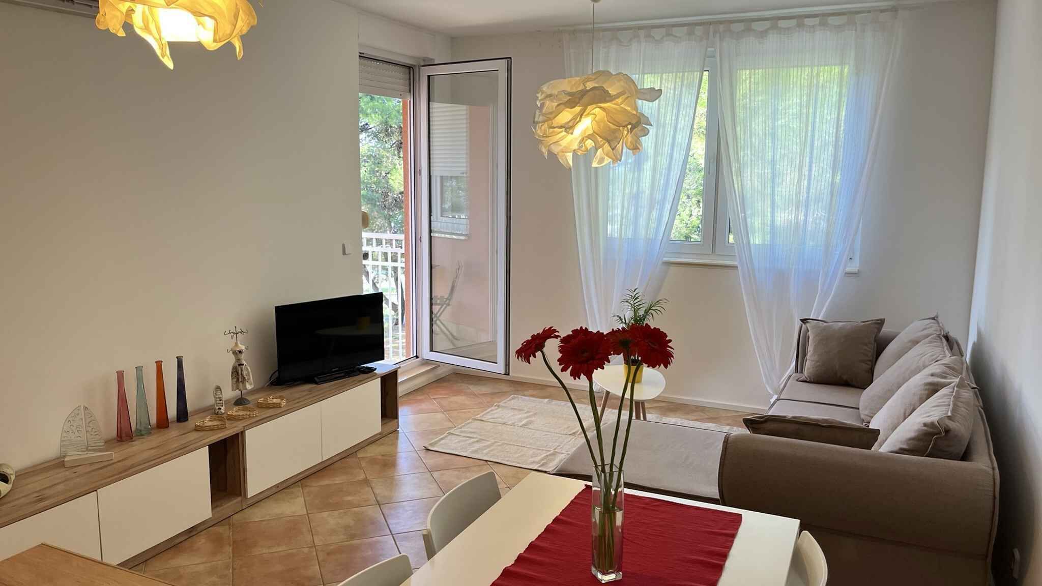 A modern living room with a television, a dining table with a red table runner, a sofa, and a vase with flowers.